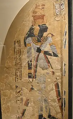 essa é uma representação da rainha Amósis-Nefertari. Ela usa um vestido longo que vai até os tornozelos, o vestido tem mangas aladas e uma gola bem grande. A rainha também usa um acessório para a cabeça e braceletes nos dois braços, todos eles em ouro.
