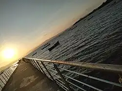 O trapiche está em perspectiva. A passarela é de concreto, com guarda-corpo metálico. No mar existem embarcações e ao fundo o sol está a se pôr.