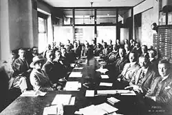 Photo of a caucus meeting showing thirty-five men and a woman, sitting around several long tables with documents laid out before them. As the space is cramped and there is not room for everyone at the table, they are seated in two rows.