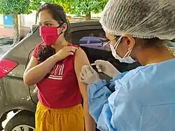 Foto de Carol Cardoso tomando uma vacina. Ela está vestida de máscara e blusa vermelha, com calças amarelas. Atrás dela, há um carro de cor metálica. Ao lado de Carol, a enfermeira que está aplicando a injeção está com uma touca para cabelo, máscara e luvas brancas, e veste-se de azul bebê. Carol é parda, de olhos e cabelos pretos, e seu cabelo é liso e longo.