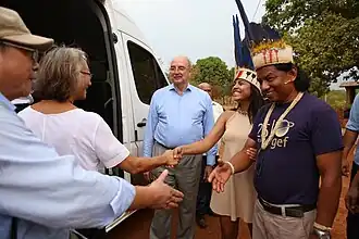 Foto com 5 pessoas em primeiro plano se cumprimentando (uma mulher indígenas com adereços típicos da cultura, um homem indígena com adereços típicos da cultura, uma mulher branca na faixa dos 60 anos e dois homens brancos entre 60 a 70 anos). Ao fundo, automóvel van branca e terreno não asfaltado com vegetação.