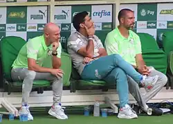 Três homens sentados, olhando para a direita. O primeiro está com o corpo inclinado pra frente, com a mão na boca. O segundo também está com a mão na boca, mas mais ereto. O terceiro está com as mãos juntas na altura dos joelhos.