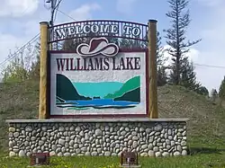 Placa de boas vindas da cidade, com a frase “Welcome to Williams Lake”, o desenho de um chapéu acima do nome da cidade e uma ilustração de lago