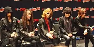 Os membros do X Japan sentados em fileira vestindo roupas pretas.