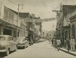 Fotografia em preto e branco de um protesto em uma rua de Maceió, durante o impeachment do governador Sebastião Muniz Falcão, na década de 1950. No alto, uma faixa com a mensagem: “IMPEACHMENT – BRUTAL VIOLAÇÃO DO DIREITO DE VOTO”. A rua de paralelepípedos é ladeada por prédios simples e carros antigos estacionados. Pessoas caminham e conversam, vestindo trajes formais, como ternos e chapéus. O ambiente reflete uma atmosfera de tensão política, com um protesto pacífico em andamento.