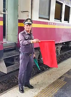 Chefe de estação ferroviária na Tailândia, com a bandeira vermelha desfraldada, significando sinal de paragem.