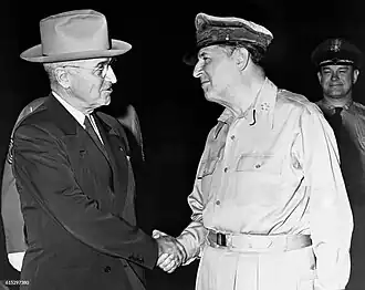 Truman in a dark suit and tie and light hat shakes hands with MacArthur, in uniform wearing a shirt but no tie and his rumpled peaked cap.