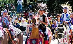 Fotografia colorida de crianças da Nação Tsuu T'ina com trajes tradicionais a cavalo em um desfile Stampede diante de uma plateia
