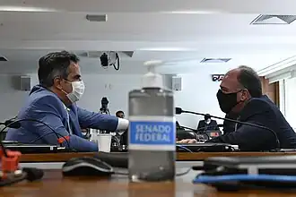 Dois homens de terno conversam ao fundo, sentados; em primeiro plano, há um álcool gel com a logomarca do Senado Federal