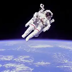 Um planeta azul com nuvens brancas irregulares, encimado por um astronauta num traje branco contra o fundo espacial negro.