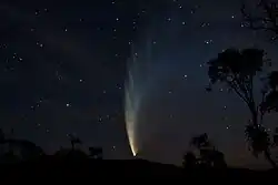 Num céu escuro, com alguma estrelas, o núcleo brilhante do cometa está próximo ao horizonte e sua calda, incialmente amarelada, se estende para o alto e para a direita mudando para a coloração azul e ficando cada vez mais extensa.