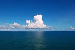 O Oceano Atlântico, azul profundo com águas calmas. No céu azul somente algumas nuvens cumulus.