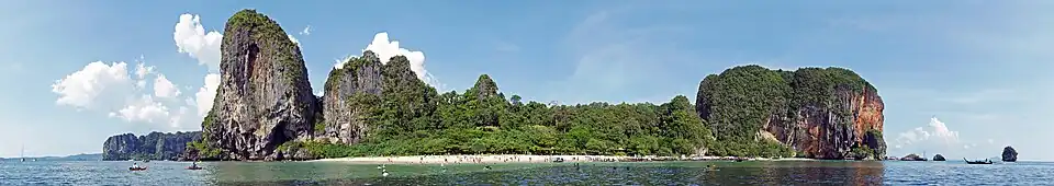 Em primeiro plano, o mar de águas azul, esverdeada. Ao centro, uma faixa de areia clara, com muitas pessoas. Alguns barcos podem ser vistos ao longe, próximo à praia. Além da praia, montanhas com paredões rochosos acinzentados a direita e à esquerda da praia, e montanhas mais suaves ao centro, cobertas pela floresta tropical.