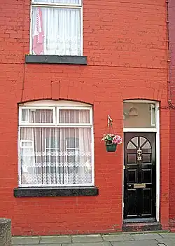 Exterior de um edifício vermelho. São visíveis uma porta preta com uma pequena janela logo acima, duas janelas maiores à esquerda da porta, uma acima da outra, e um vaso de flores entre a porta e a janela maior mais de baixo.