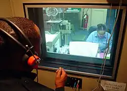 homem usando fones de ouvido fazendo um teste audiológico dentro da uma cabine, com uma profissional de audiologia realizando o teste