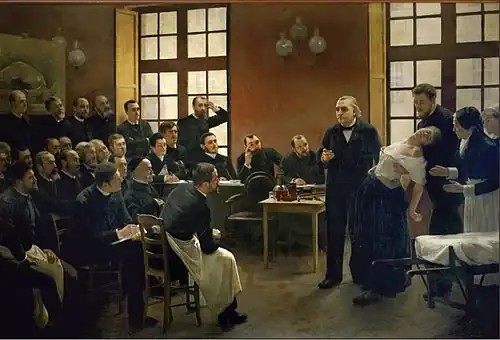 Pintura de uma aula médica do século XIX. Ao fundo, uma mulher desmaia nos braços de um homem que se encontra logo atrás dela, enquanto outra mulher, aparentemente enfermeira, se prepara para ajudar. Um homem idoso, o professor, gesticula como se estivesse fazendo um apontamento. Cerca de duas dezenas de estudantes do sexo masculino observam a cena.