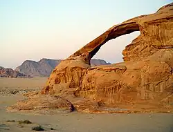 Arco natural inclinado, semelhante a um puxador de gaveta. formando um "buraco" na colina, é possível ver o deserto montanhoso em que se encontra, ao fundo através do arco.