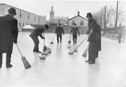 Um grupo de homens, vestidos com roupas de inverno e usando chapéus, participam de uma partida de curling. Um deles varre o gelo, enquanto os outros, também com vassouras na mão, observam.