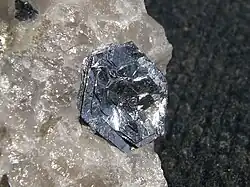 Cristais lustrosos, prateados, planos e hexagonais em camadas quase paralelas dispostas em formato parecido com o de uma flor em um pedaço cristalino de quartzo.