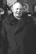 A man wearing a long coat, smiling, in front of a group of people and the Soviet flag