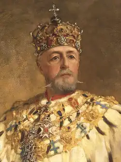 Um homem idoso e barbudo em trajes ornamentados, usando uma coroa adornada com joias encimada por uma cruz, olha diretamente para fora da imagem.