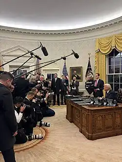 Trump, seated at the Resolute Desk in the White House, looks onward to a crowd of reporters with cameras and boom microphones