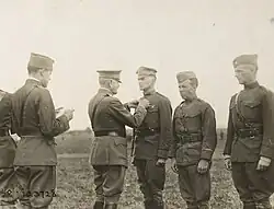 Three men in uniform are standing side by side. The one on the left is wearing a peaked "crush cap" and standing smartly at attention, while the two on the right wear garrison caps and are slouching. A man in a peaked cap and Sam Browne belt is pinning something on the chest of the first man. Behind him stands another man in a garrison cap who is reading a document in his hands.