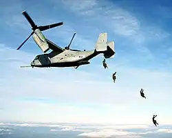 foto colorida de quatro paraquedistas saltando da rampa aberta de um MV-22 Osprey em vôo