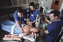 estudantes com uniformes azul escuro em torno de um boneco de plástico de tamanho humano em uma maca com equipamentos de medicina