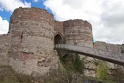 Duas torres redondas de pedra amarelo claro na parte inferior e pedra laranja escura no topo de cada lado de uma entrada arqueada. Uma ponte leva da entrada para permitir o acesso.