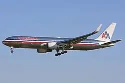 Side quarter view of twin-engine jetliner in flight, with extended gear; bare metal with red, white, and blue cheatlines, as well as two "A"s and a stylized eagle on the tail