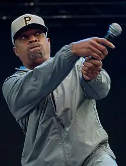 Rapper Chuck D fazendo o movimento de um taco de baseball com o microfone enquanto performa. Ele usa um boné do time de baseball Pittsburgh Pirates.
