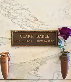 Cripta de mármore com placa de latão inscrita: Clark Gable, 1º de fevereiro de 1901 16 de novembro de 1960. Dois vasos são anexados ao  cripta, uma contém uma rosa roxa.