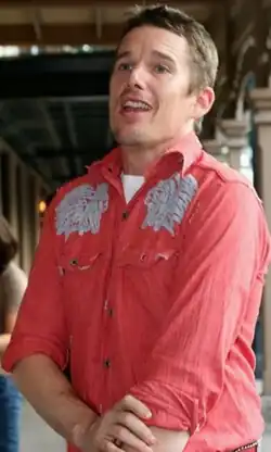 A caucasian male with light brown hair and light brown sttuble is facing to the left. He is wearing a red shirt, with a white t-shirt visible underneath. The red shirt has a white pattern on the shoulders.