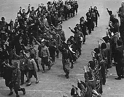 Several lines of school children march diagonally from top right to bottom left. Each carries a bag or bundle and each raises their right arm in the air in a salute. Adults stand in a line across the bottom right hand corner making the same gesture.