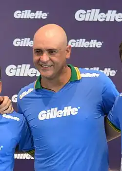 Um homem branco, careca, sorrindo e não olhando para a câmera. Ele veste uma camisa azul escrito "Gillette".