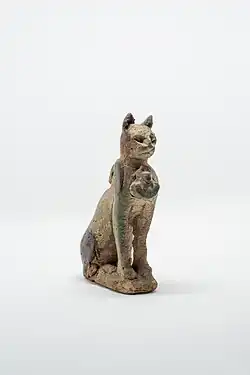 Representação de um gato sentado sobre as patas traseiras com a cauda junto ao lado direito do seu corpo. No peito um aegis com cabeça de uma leoa com o disco solar. Possui um anel para sustentação nas costas. O revestimento vítreo desapareceu quase completamente. Item da Coleção Egípcia da Casa Museu Eva Klabin