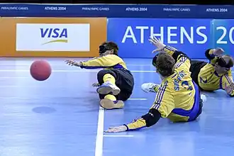 Três atletas, usando vendas, estão deitados na quadra, durante uma partida de goalball.