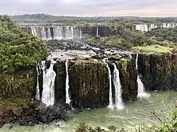 Conjunto de cachoeiras caindo sobre paredões de rocha basáltica escura, em dois degraus sequenciais, rodeado por vegetação de floresta.