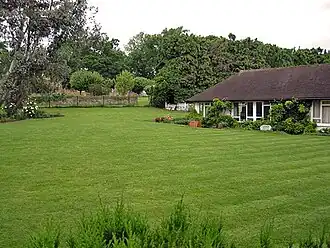 Uma casa branca com um telhado preto ao lado de uma grande jardim bem cuidado. Há várias árvores no fundo da imagem, atrás da casa.