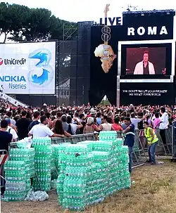 A imagem mostra parte do palco com um grande público. Destaca-se o mapa da África no lado esquerdo e a  imagem de um telão com a apresentação em curso. Há a presença também de vários montes de garrafas de água no lado direito.