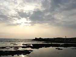 Imagem do mar mediterrâneo com algumas nuvens no céu. Há uma formação rochosa no lado direito e algumas pessoas andam em cima dela.