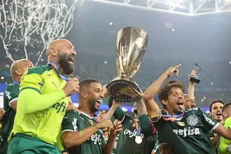 "Vários homens felizes, sorrindo, comemorando e pulando. No centro uma taça de ouro é erguida por um jogador."