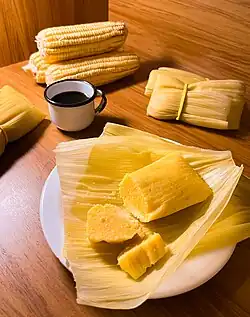 Fotografia de pamonhas embaladas em palha de milho servidas com café e espigas de milho verde. No centro da imagem há uma pamonha desembalada e cortada sobre um prato