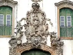 Armas em formato de cartelas de estilo rococó, com a bordadura de castelos substituída por estrelas representando as capitanias do Brasil. Conjunto escultórico d'o Aleijadinho na Igreja de São Francisco de Assis de Ouro Preto, século XVIII