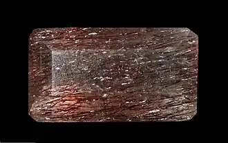 Mineral de formato retangular, com quatro lados e de cor avermelhada com algumas manchas mais escuras.