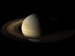 Saturno no centro da imagem, com sua metade esquerda iluminada, com coloração bege. Os anéis aparecem com formato elíptico, devido à visão oblíqua, marrons.