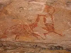 Pintura rupestre avermelhada sobre uma rocha mais clara, parece representar um humano interagindo com um grande animal