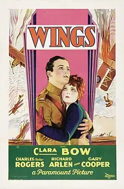 Cartaz promocional de um filme. Ele se concentra em duas pessoas. O homem está vestindo um terno de aviador e a mulher está vestindo uma jaqueta azul e luvas vermelhas.