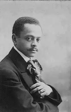 Fotografia em preto e branco de um homem negro com bigode e cabelo curto, vestindo terno e gravata, olhando diretamente para a câmera.